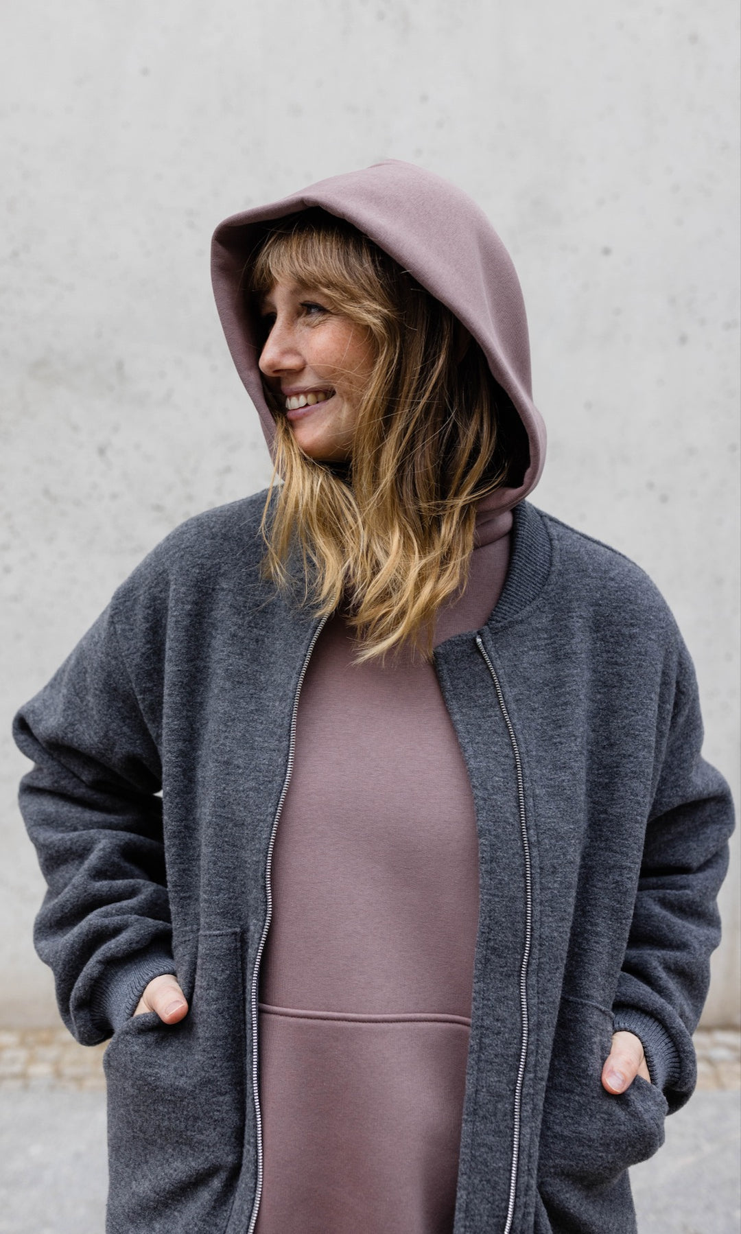 Still Hoodie in aubergine rot stehend  im Herbst mit schwarzer Hose und grauer Jacke vor grauer Wand Kapuze auf Kopf Hände in Tasche der Jacke von vorne Blick seitlich lächelnd
