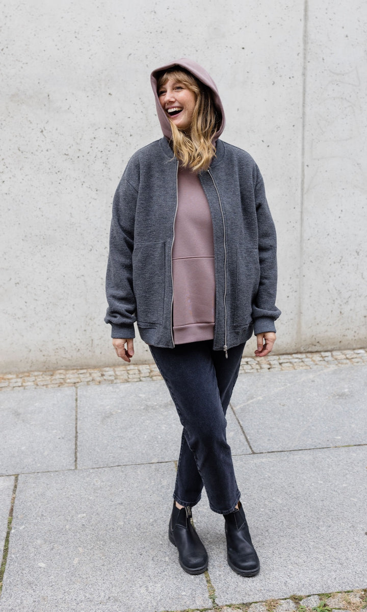 Still Hoodie in aubergine rot stehend im Winter mit schwarzer Hose und Stiefeln und grauer Jacke vor grauer Wand Kapuze auf Kopf von vorne Blick seitlich lachend