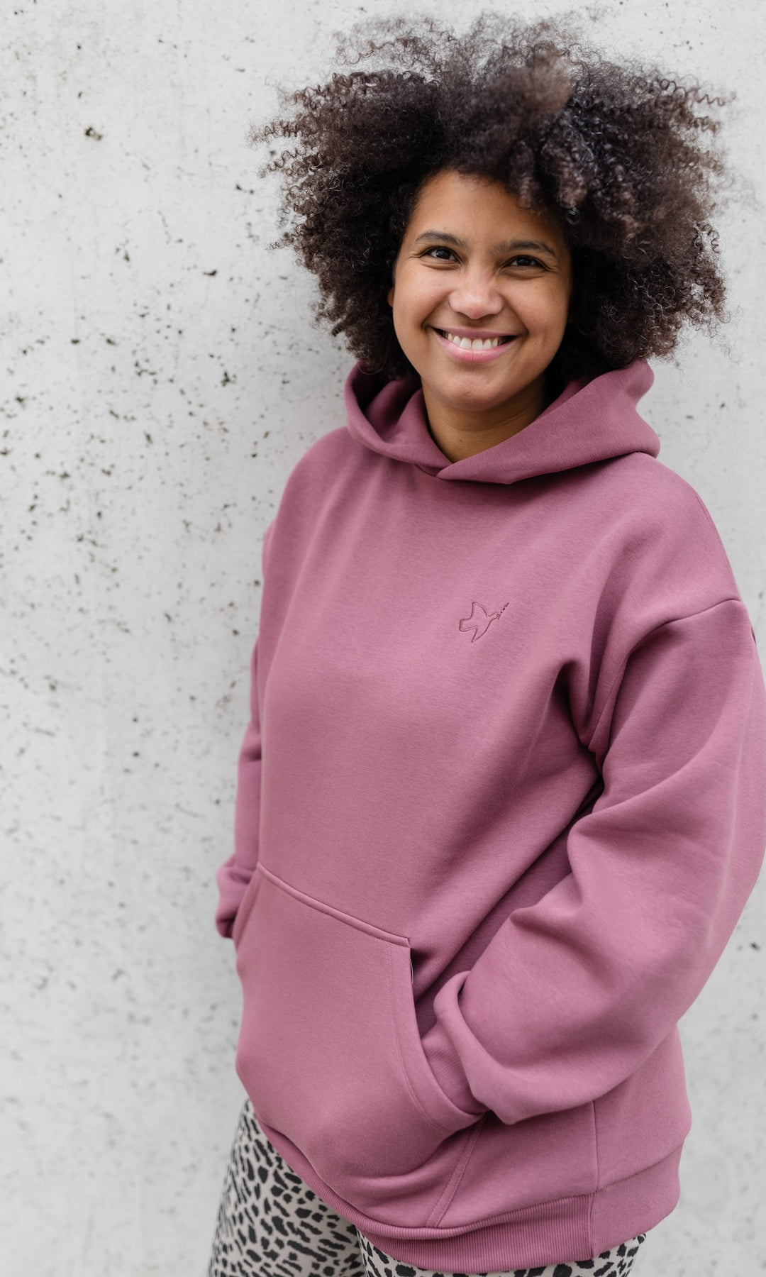 Stillhoodie in feige rosa vor grauer Wand kombiniert mit Leo Leggings seitlich Blick gerade lächelnd