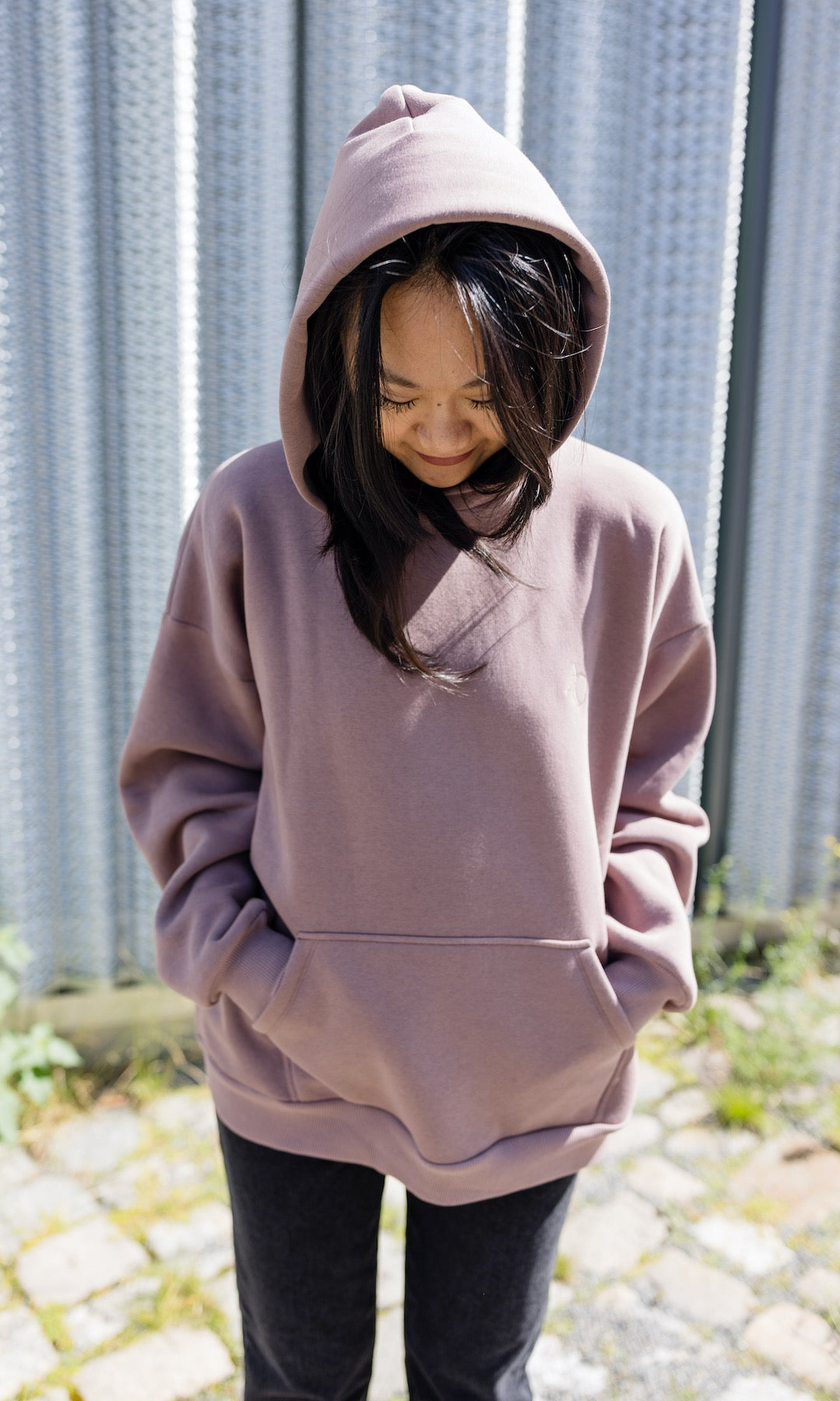 Still Kapuzenpullover in aubergine rot stehend mit schwarzer Hose Kapuze auf Kopf vor grauer Wand Nahaufnahme von vorne Blick nach unten