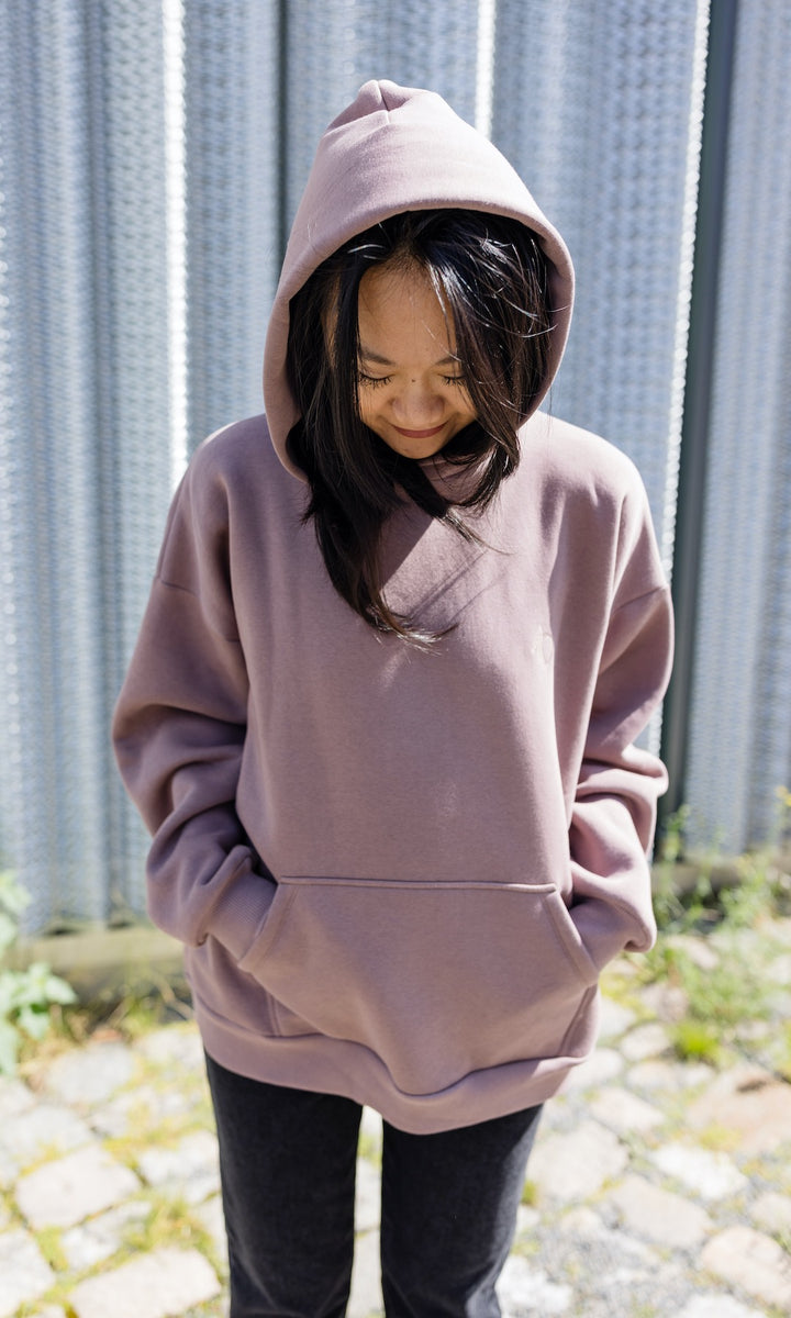 Still Kapuzenpullover in aubergine rot stehend mit schwarzer Hose Kapuze auf Kopf vor grauer Wand Nahaufnahme von vorne Blick nach unten