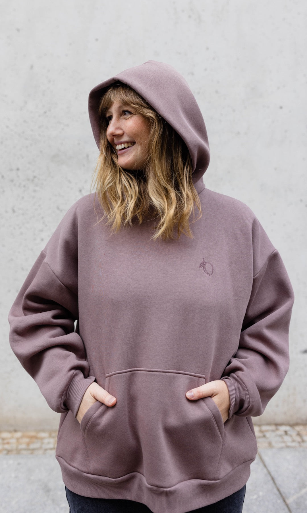 Still Kapuzenpullover in aubergine rot stehend mit schwarzer Hose vor grauer Wand Hände in Tasche von vorne Blick seitlich lächelnd