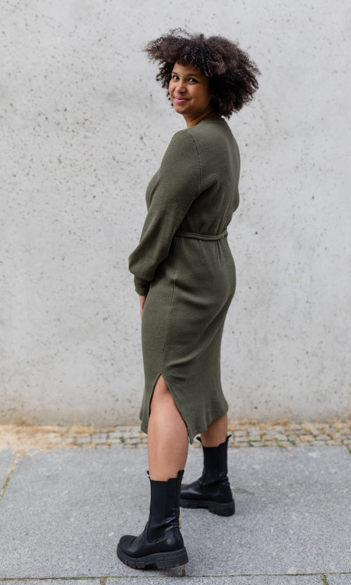 Still Strickkleid Jules in grün olive mit Stiefeln seitlich vor heller Wand