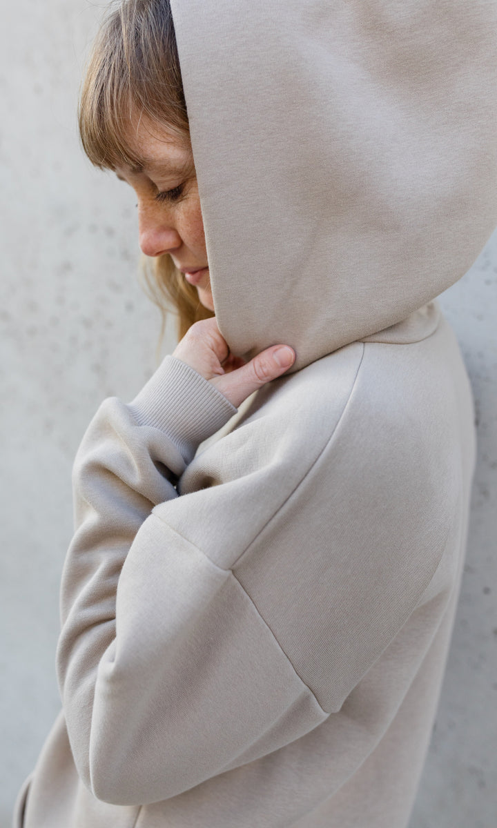 Stillhoodie in sesam weiß stehend seitlich vor grauer Wand Kapuze auf Kopf seitlich Blick nach unten 