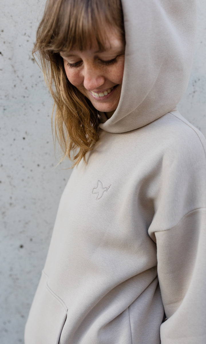 Stillhoodie in sesam weiß stehend seitlich vor grauer Wand Kapuze auf Kopf seitlich Blick nach unten lachend