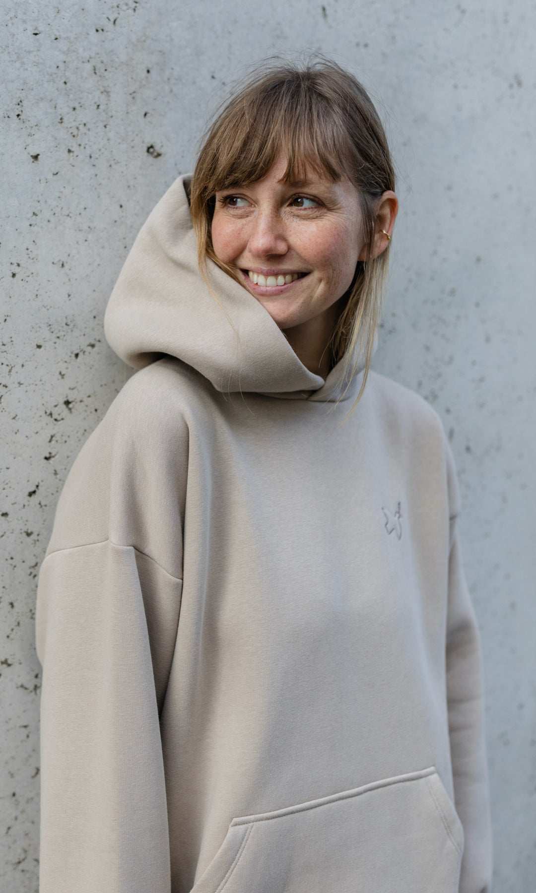 Stillhoodie in sesam weiß stehend vor grauer Wand seitlich Blick seitlich lächelnd