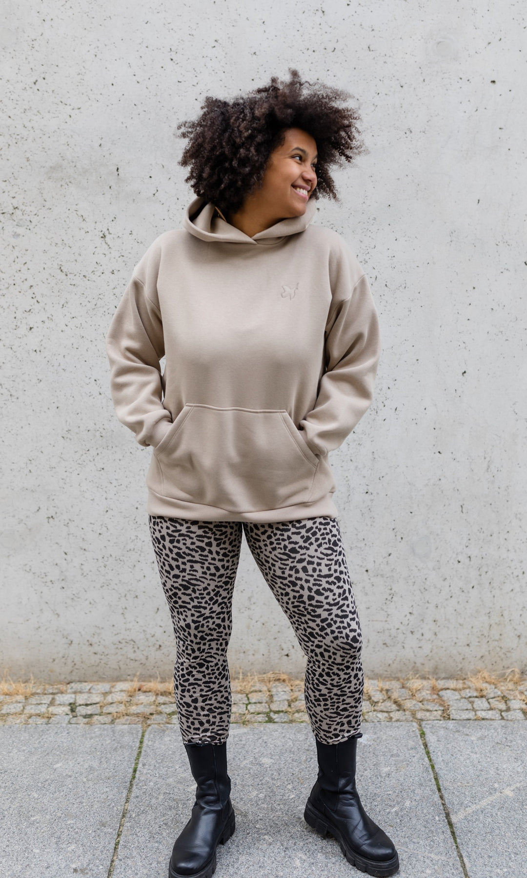 Stillhoodie in sesam weiß stehend mit Leo Leggings und schwarzen Stiefeln vor grauer Wand von vorne Blick seitlich lachend
