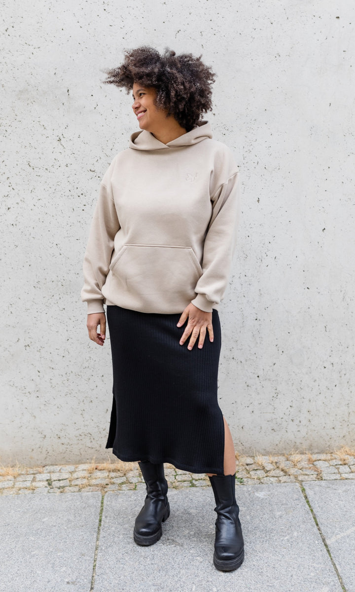 Stillhoodie in sesam weiß stehend mit schwarzem Rock und schwarzen Stiefeln vor grauer Wand von vorne Blick seitlich lachend