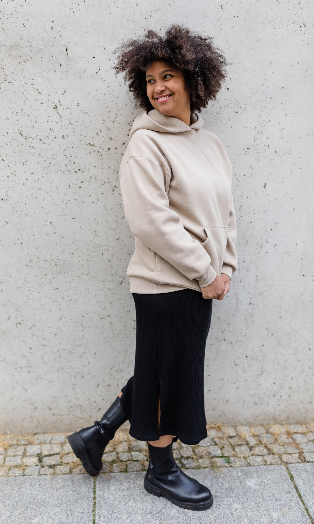 Stillhoodie in sesam weiß seitlich stehend an grauer Wand mit schwarzem Rock und Stiefeln von vorne Blick seitlich