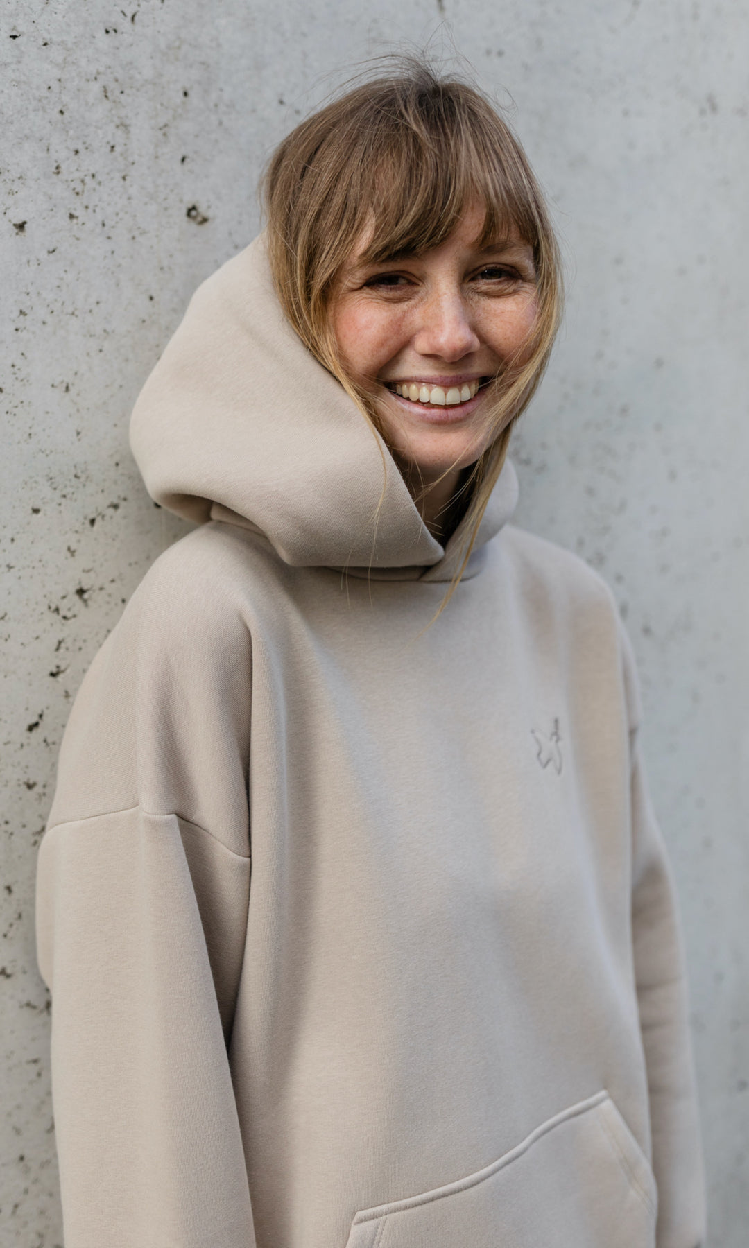 Stillhoodie in sesam weiß stehend vor grauer Wand lachend