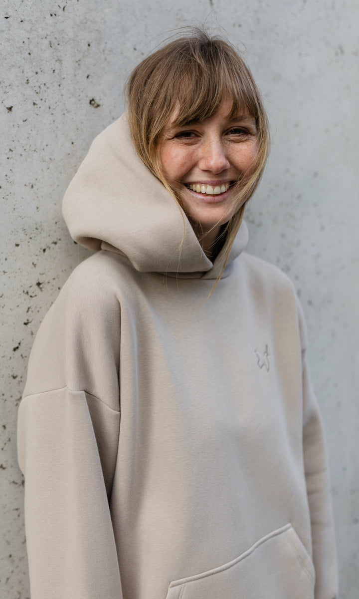 Stillhoodie in sesam weiß stehend vor grauer Wand lachend