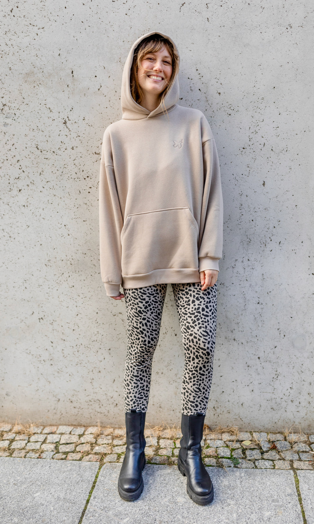 Stillhoodie in sesam weiß stehend im Herbst mit Leo Leggings und schwarzen Stiefeln vor grauer Wand Kapuze auf Kopf von vorne Blick in die Kamera