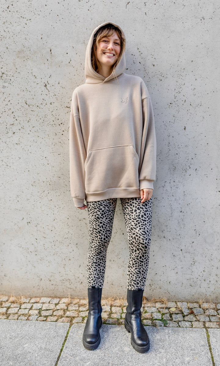 Stillhoodie in sesam weiß stehend im Herbst mit Leo Leggings und schwarzen Stiefeln vor grauer Wand Kapuze auf Kopf von vorne Blick in die Kamera