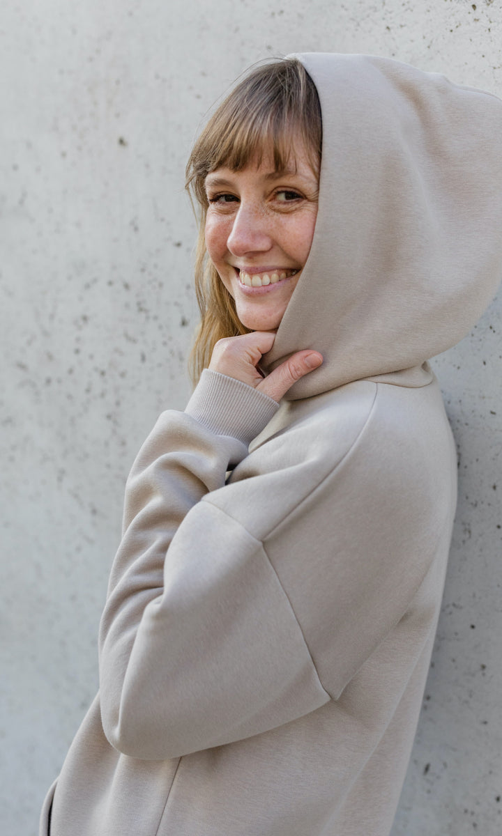 Stillhoodie in sesam weiß stehend im Herbst angelehnt an graue Wand Kapuze auf Kopf seitlich Blick in die Kamera