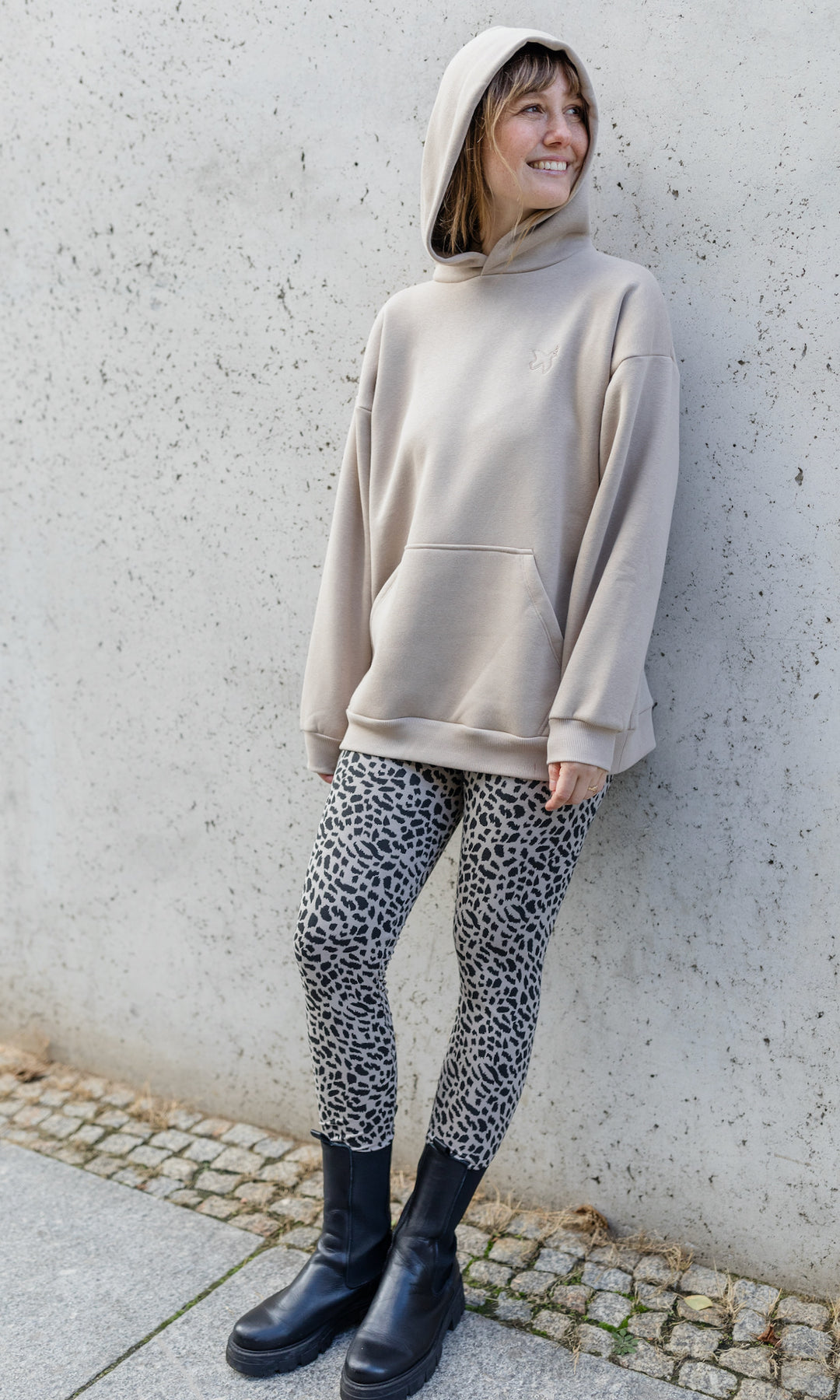 Stillhoodie in sesam weiß stehend im Herbst mit Leo Leggings und schwarzen Stiefeln vor grauer Wand Kapuze auf Kopf seitlich Blick seitlich lächelnd