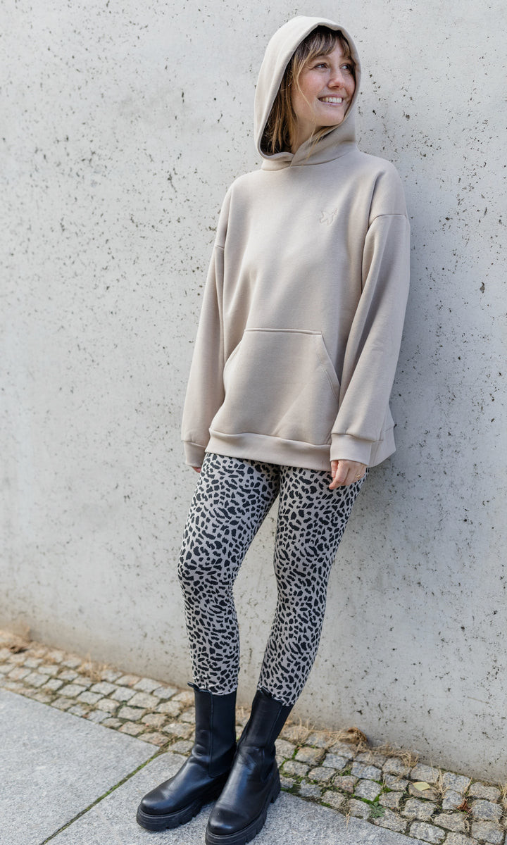 Stillhoodie in sesam weiß stehend im Herbst mit Leo Leggings und schwarzen Stiefeln vor grauer Wand Kapuze auf Kopf seitlich Blick seitlich lächelnd