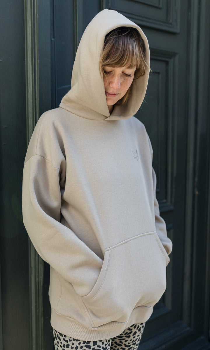 Stillhoodie in sesam weiß stehend im Winter mit Leo Leggings und schwarzen Stiefeln vor grauer Wand Kapuze auf Kopf Hände in Taschen seitlich Blick nach unten