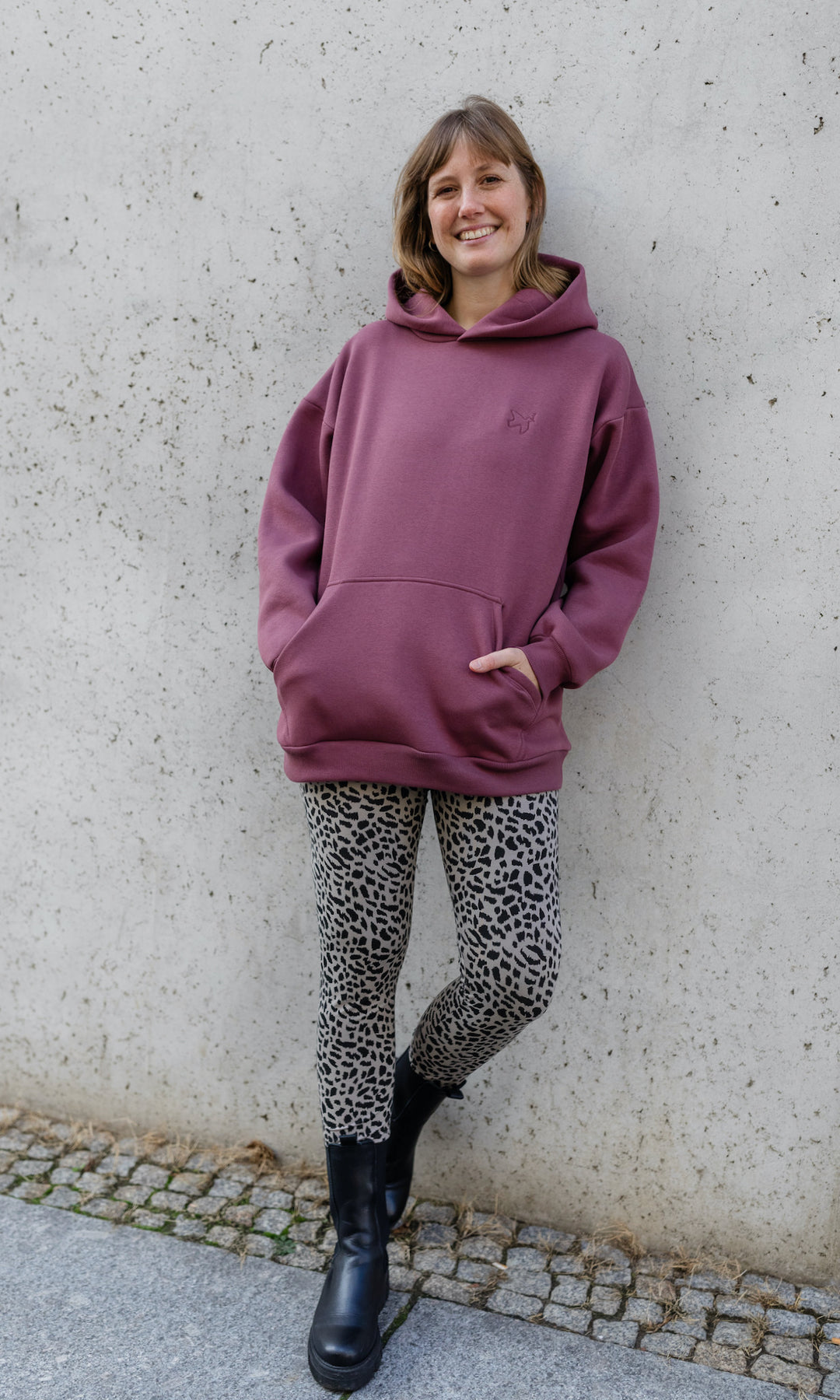 Stillhoodie in feige rosa vor grauer Wand kombiniert mit Leo Leggings und schwarzen Stiefeln von vorne Blick gerade lächelnd