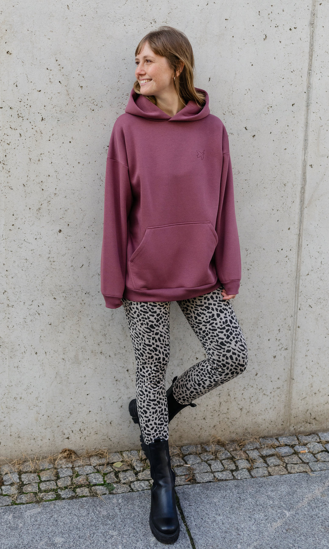 Stillhoodie in feige rosa angelehnt an graue Wand kombiniert mit Leo Leggings seitlich Blick seitlich lächelnd