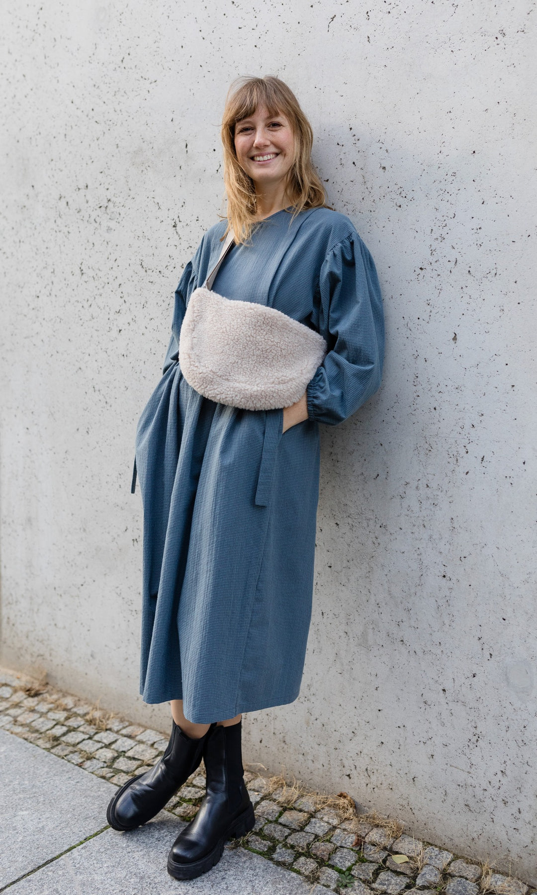 Langes Stillkleid Anouk in karo blau mit Tasche und Boots angelehnt an Wand