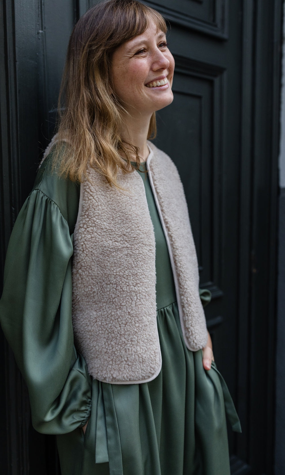 Stillkleid Shania im Herbst in olive grün mit Schafwoll-Weste angelehnt an Tür seitlich