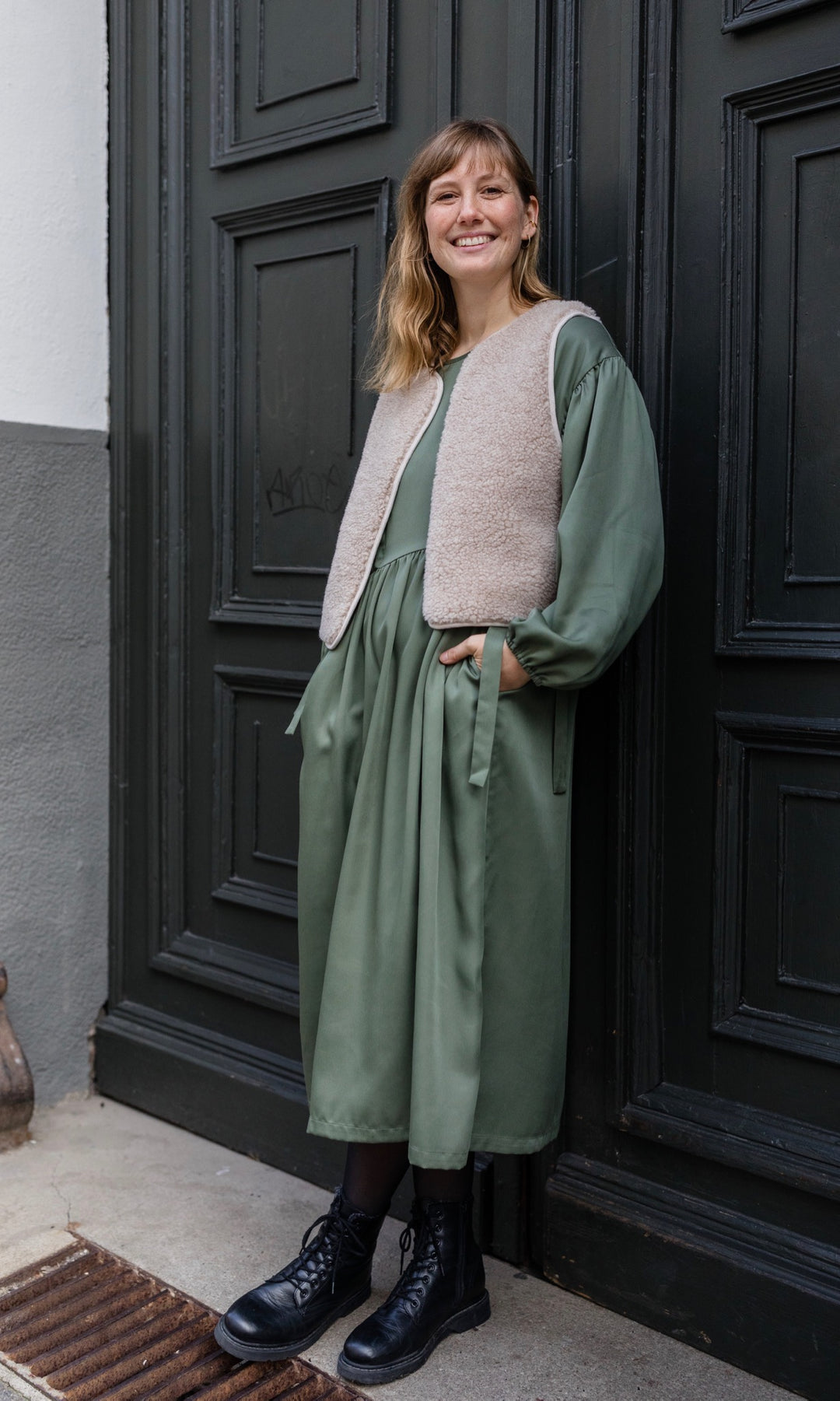 Stillkleid Shania im Herbst in olive grün mit Schafwoll-Weste seitlich angelehnt an Tür