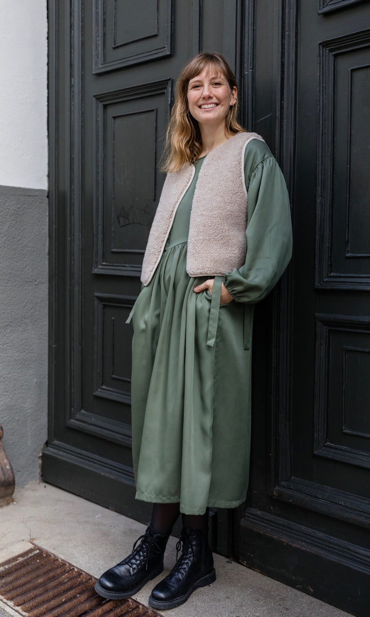 Stillkleid Shania im Herbst in olive grün mit Schafwoll-Weste seitlich angelehnt an Tür