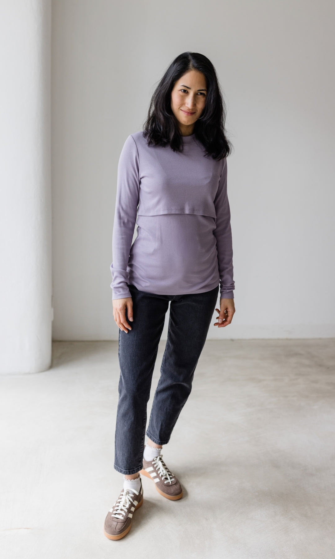 Stillshirt lang mit Rollkragen in flieder kombiniert mit dunkelblauer Jeans und Sneakers vor grauer Wand von vorne Blick gerade