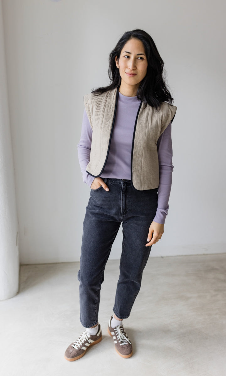 Stillshirt lang mit Rollkragen in flieder kombiniert mit dunkelblauer Jeans, heller Steppweste und Sneakers vor grauer Wand von vorne Blick gerade