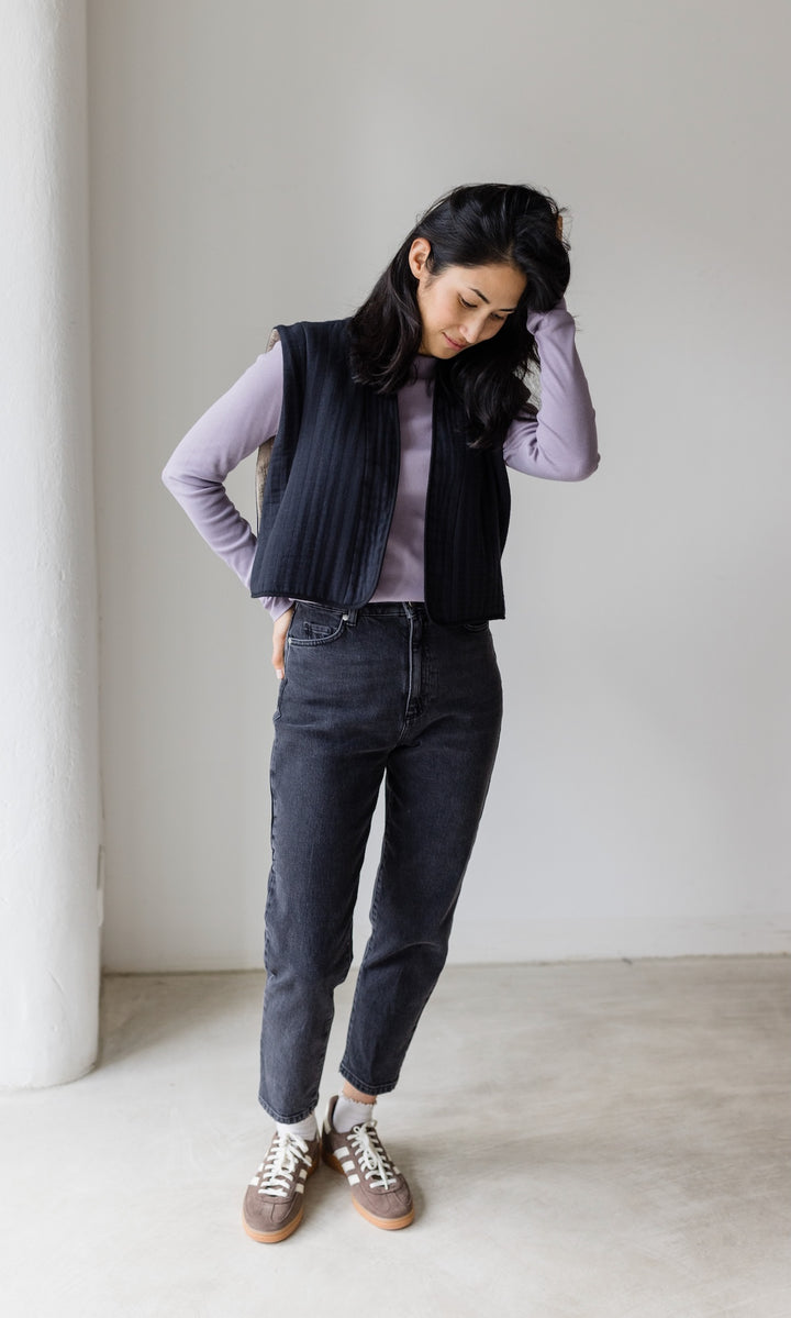 Stillshirt lang mit Rollkragen in flieder kombiniert mit dunkelblauer Jeans, dunkler Steppweste und Sneakers vor grauer Wand von vorne Blick nach unten