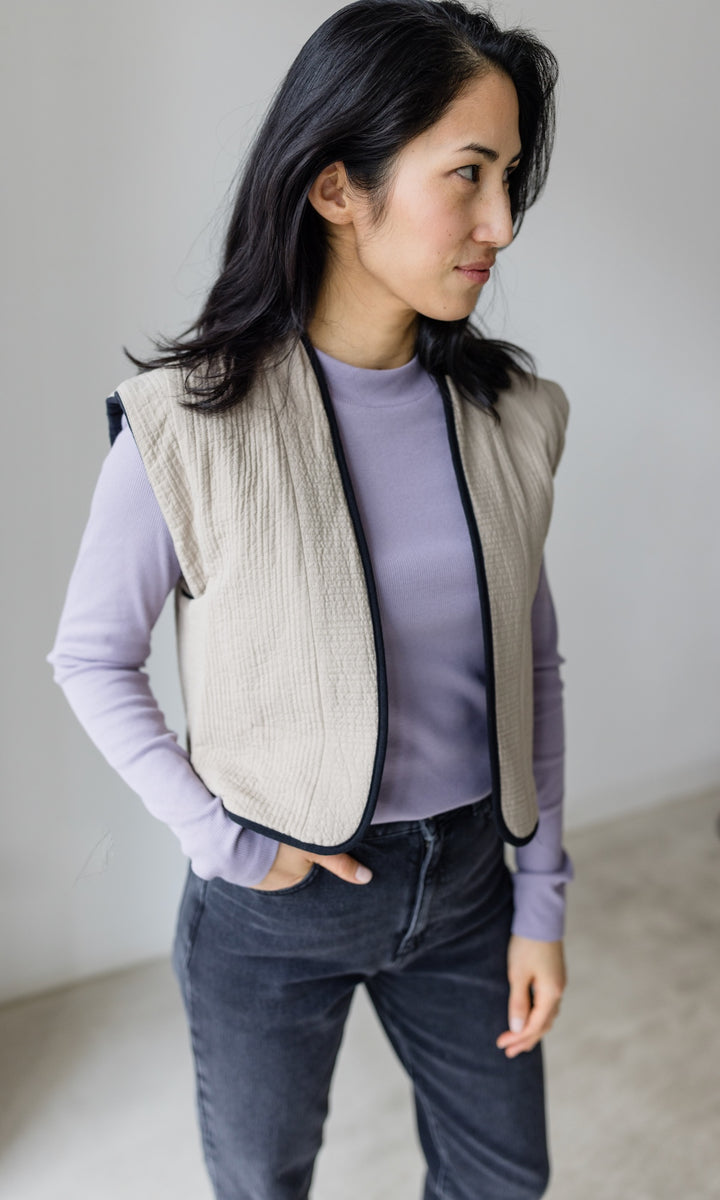 Stillshirt lang mit Rollkragen in flieder kombiniert mit dunkelblauer Jeans, heller Steppweste und Sneakers vor grauer Wand seitlich Blick gerade