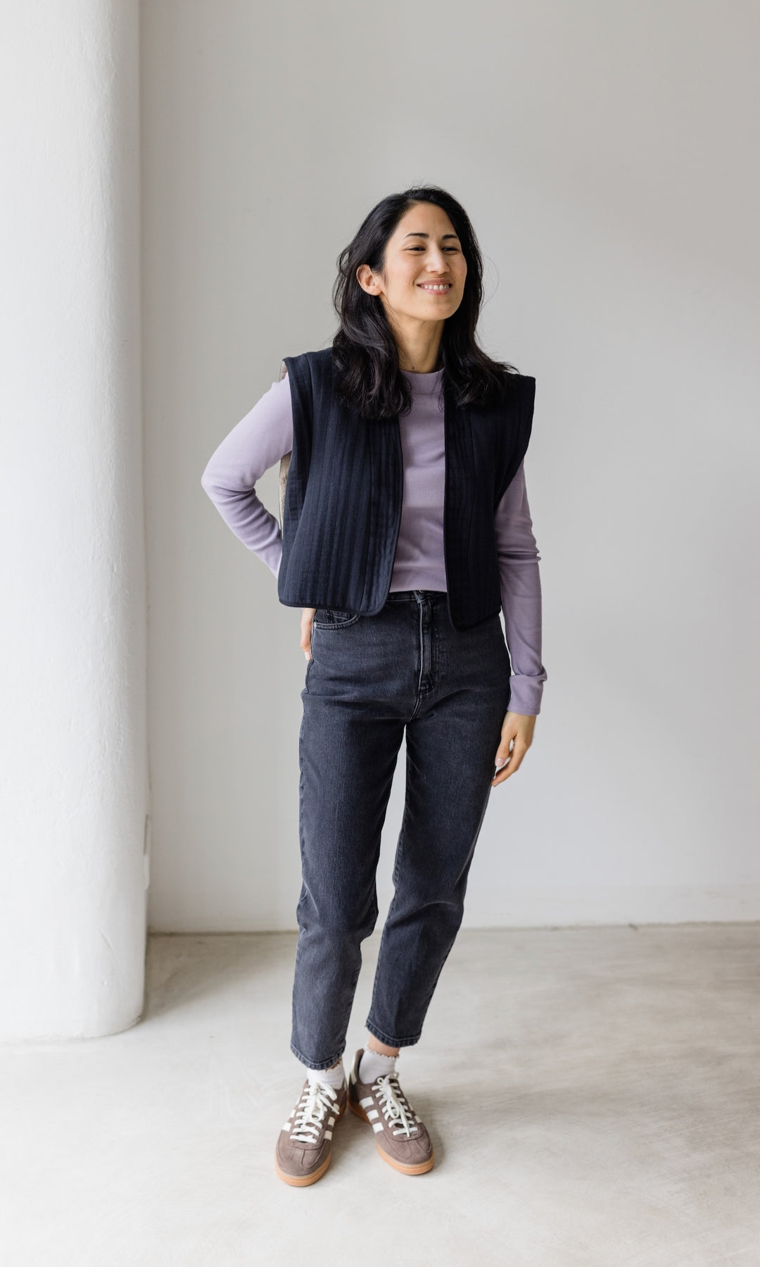 Stillshirt lang mit Rollkragen in flieder kombiniert mit dunkelblauer Jeans, dunkler Steppweste und Sneakers vor grauer Wand von vorne Blick lachend