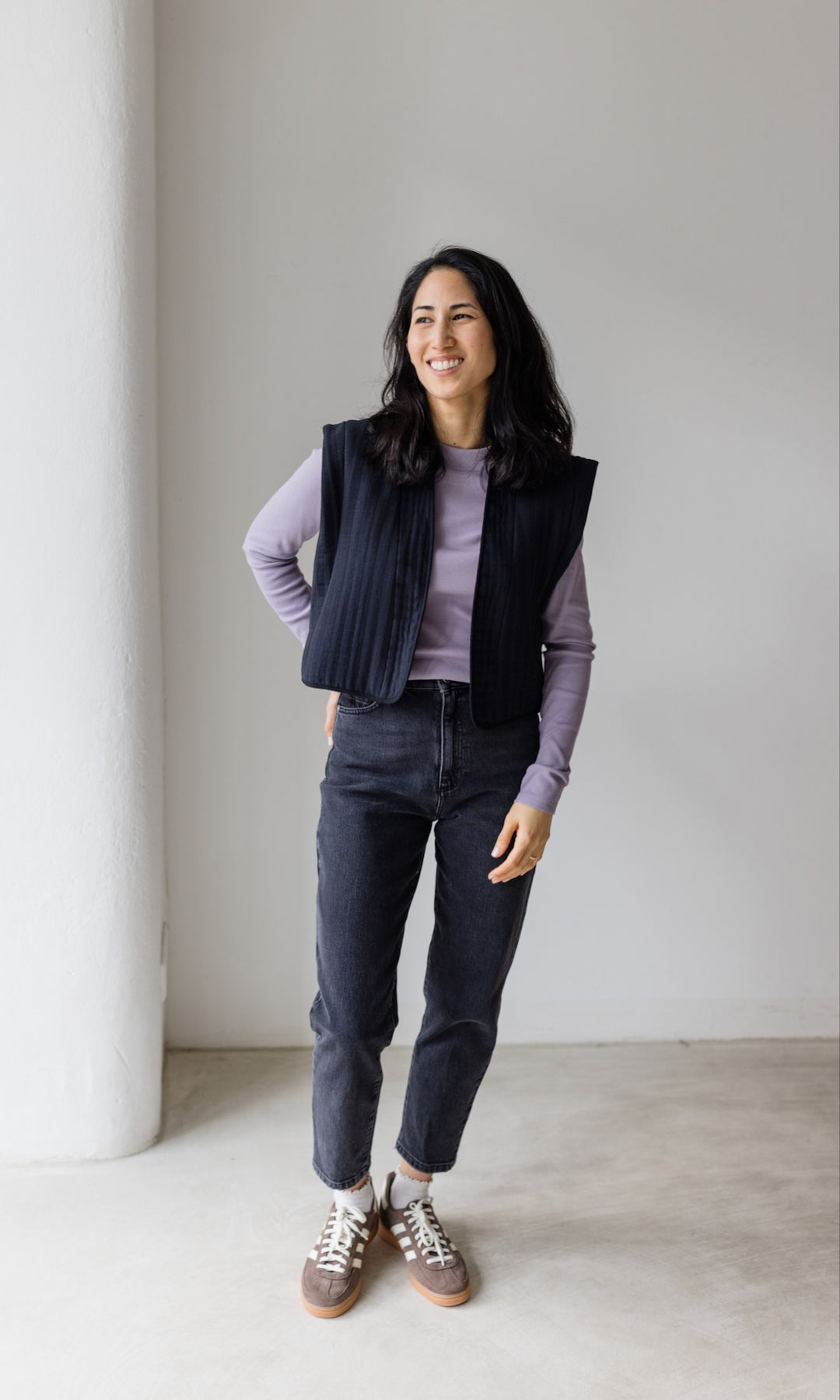 Stillshirt lang mit Rollkragen in flieder kombiniert mit dunkelblauer Jeans, dunkler Steppweste und Sneakers vor grauer Wand von vorne Blick seitlich lachend