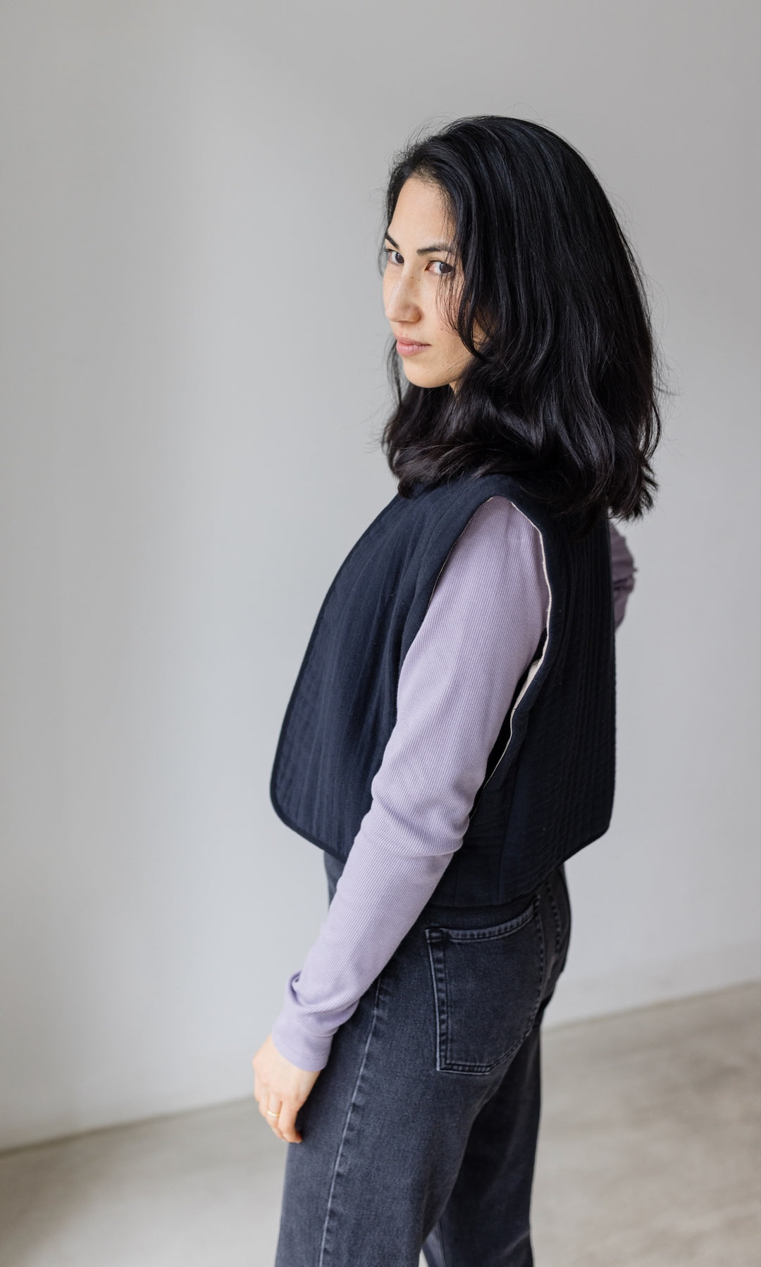 Stillshirt lang mit Rollkragen in flieder kombiniert mit dunkelblauer Jeans, dunkler Steppweste und Sneakers vor grauer Wand seitlich Blick seitlich