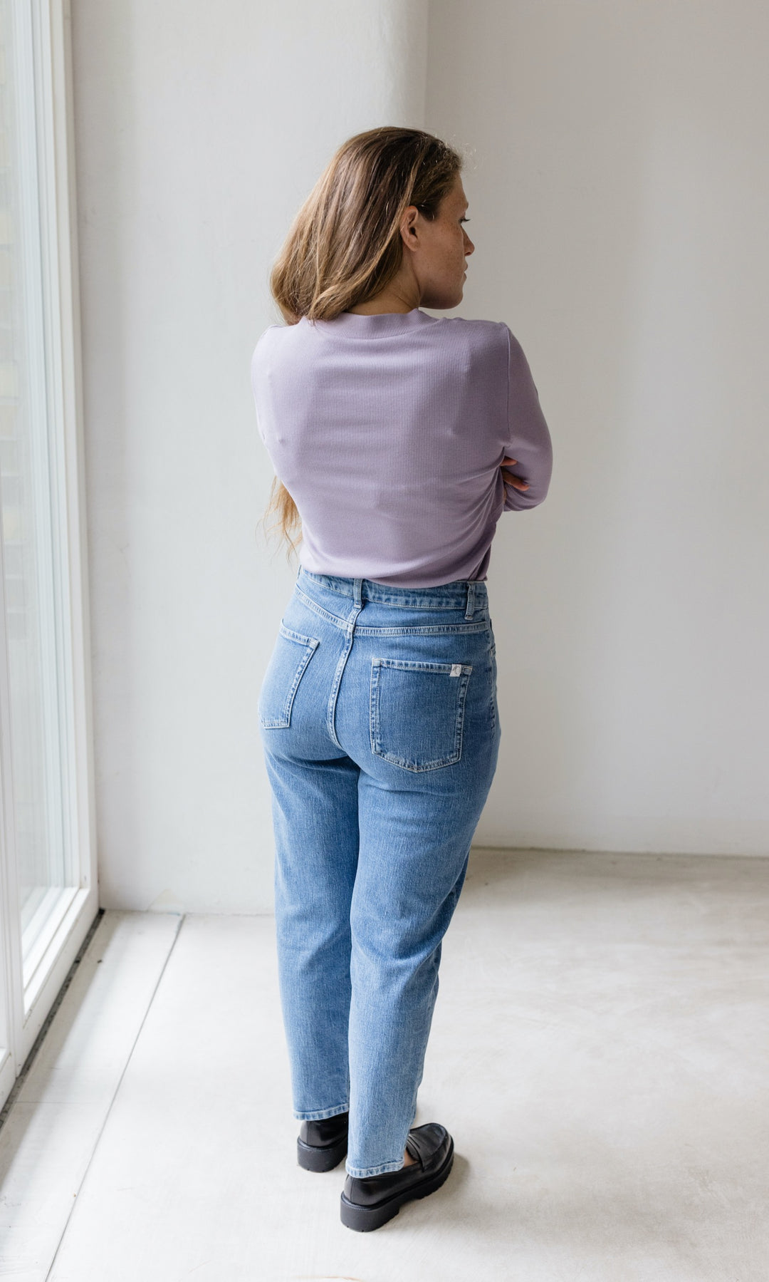 Stillshirt lang mit Rollkragen in flieder kombiniert mit hellblauer Jeans vor grauer Wand von hinten Blick seitlich