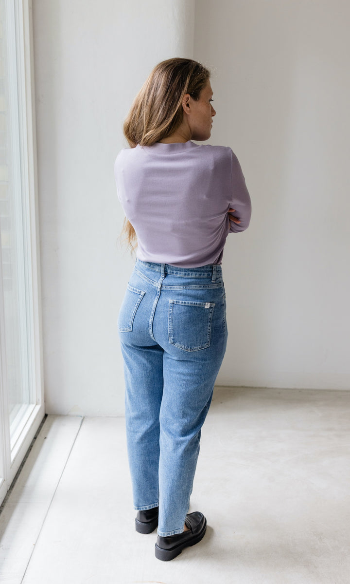 Stillshirt lang mit Rollkragen in flieder kombiniert mit hellblauer Jeans vor grauer Wand von hinten Blick seitlich