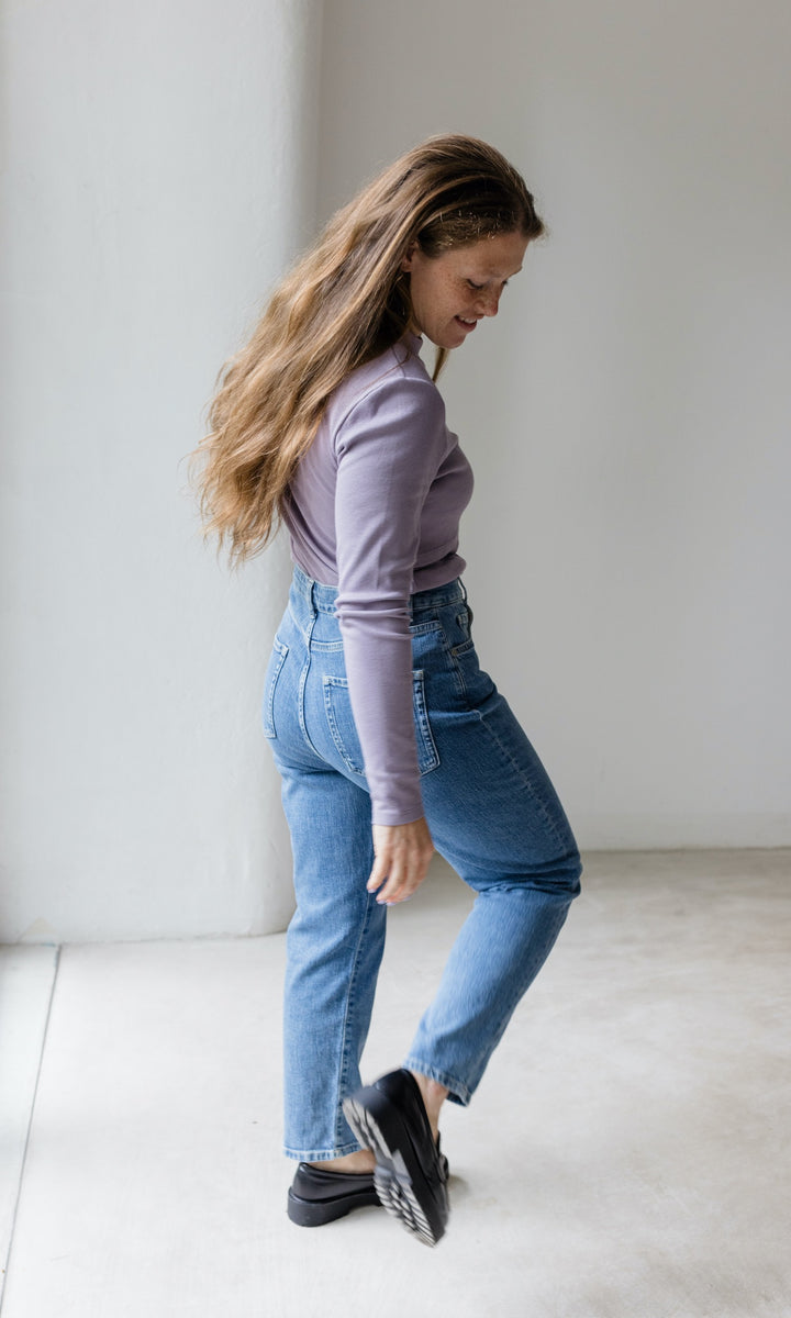 Stillshirt lang mit Rollkragen in flieder kombiniert mit hellblauer Jeans vor grauer Wand von hinten Blick nach unten