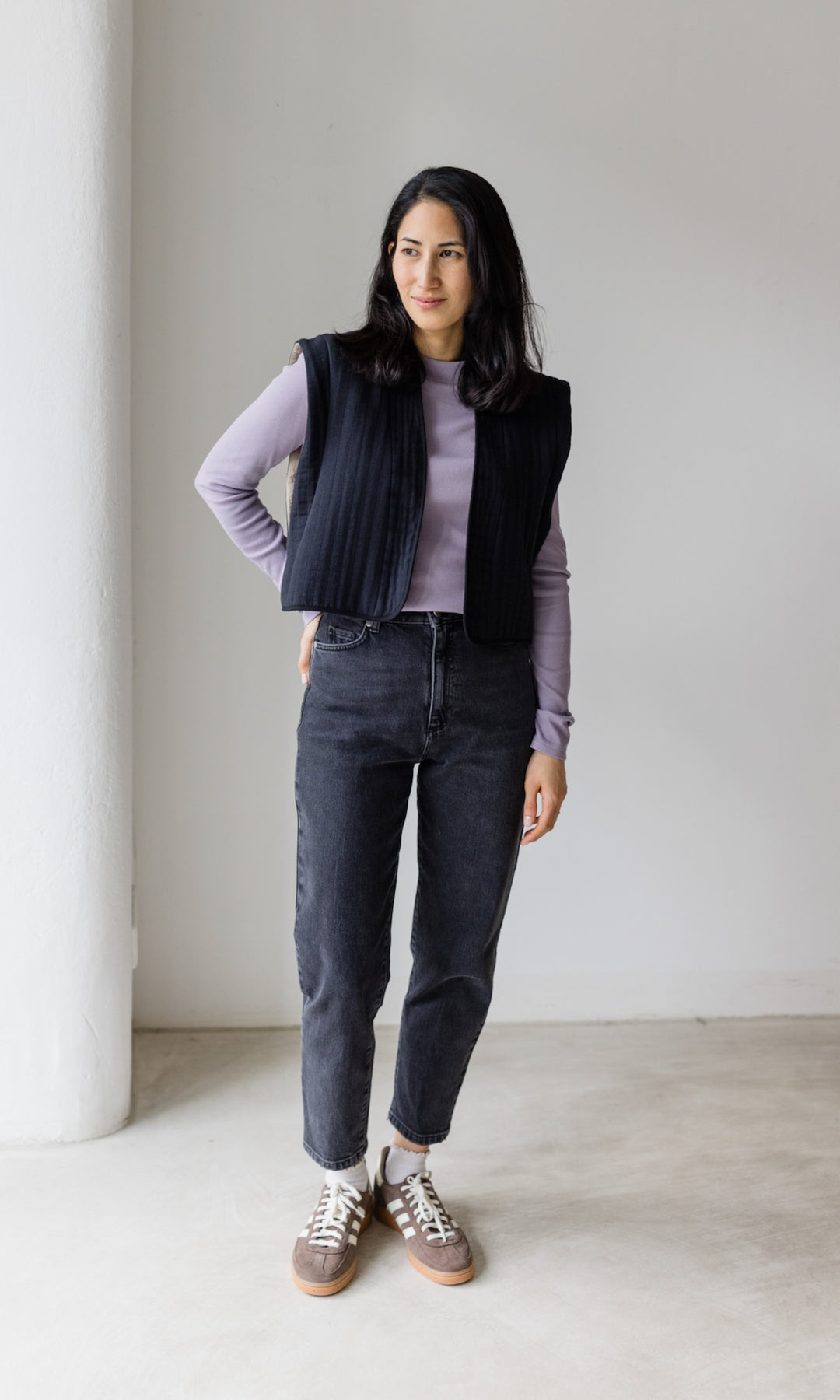 Stillshirt lang mit Rollkragen in flieder kombiniert mit dunkelblauer Jeans, dunkeler Steppweste und Sneakers vor grauer Wand von vorne Blick seitlich