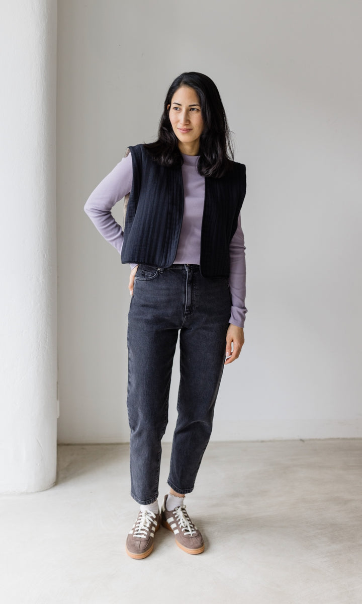 Stillshirt lang mit Rollkragen in flieder kombiniert mit dunkelblauer Jeans, dunkeler Steppweste und Sneakers vor grauer Wand von vorne Blick seitlich