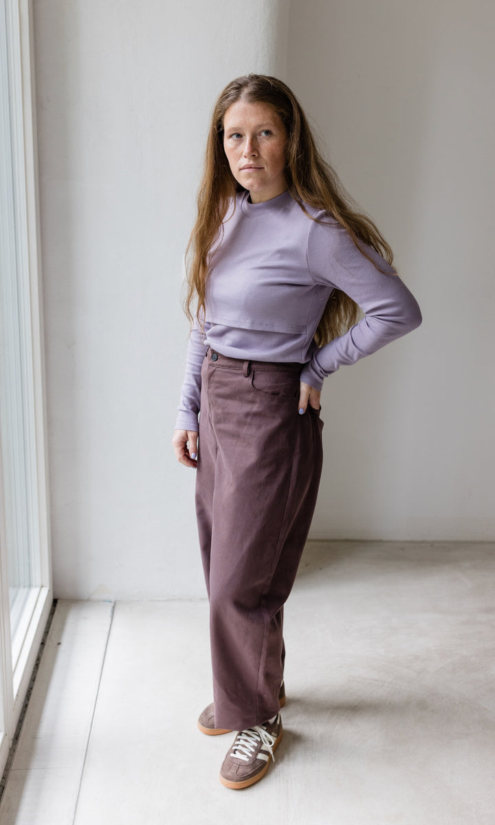 Stillshirt lang mit Rollkragen in flieder kombiniert mit roter, lila Hose und Sneakers vor grauer Wand seitlich Blick gerade