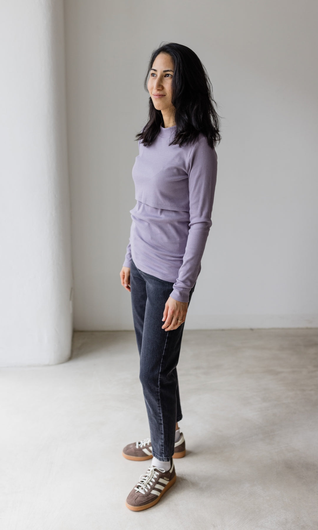 Stillshirt lang mit Rollkragen in flieder kombiniert mit dunkelblauer Jeans und Sneakers vor grauer Wand seitlich Blick gerade