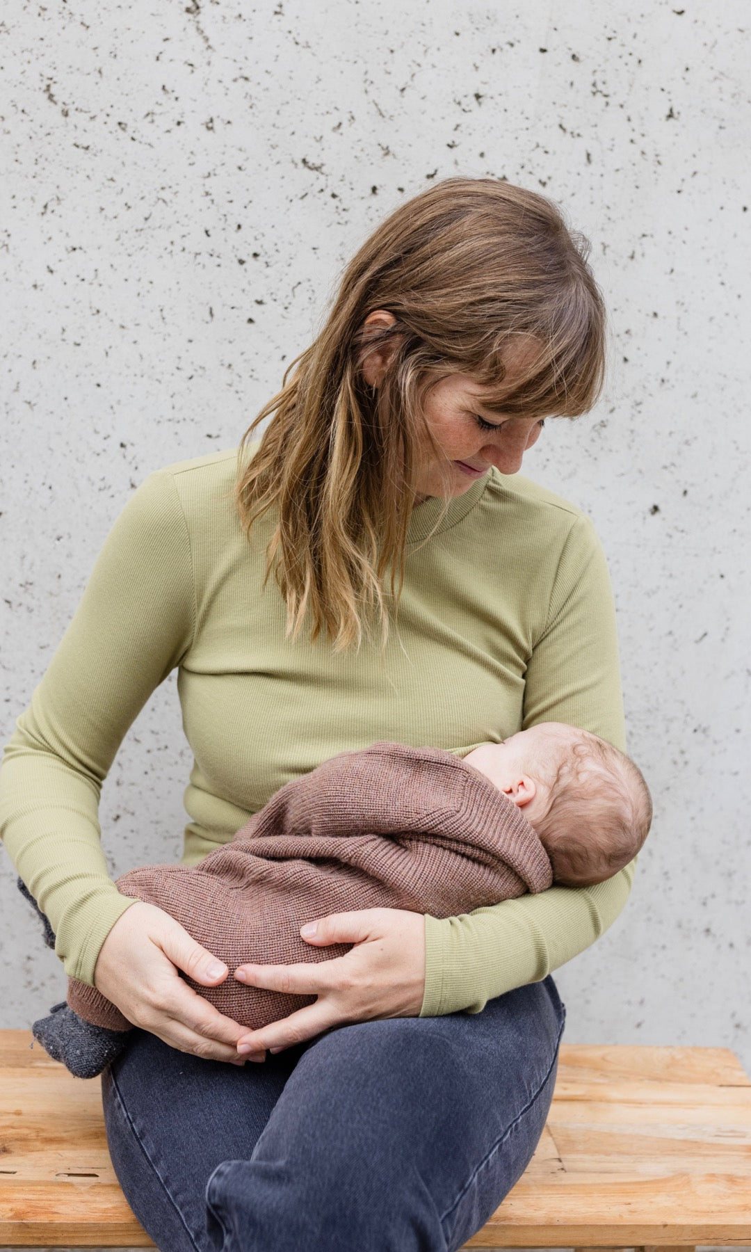 Stillshirt langarm Cléo mit Rollkragen in olive grün kombiniert mit dunkelblauer Jeans sitzend auf Bank mit Baby auf Arm beim Stillen