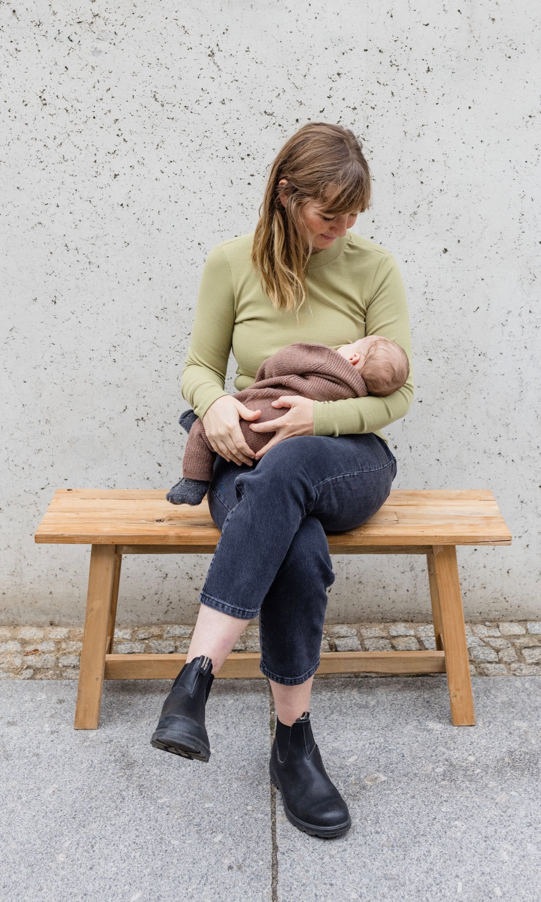 Stillshirt langarm Cléo mit Rollkragen in olive grün kombiniert mit dunkelblauer Jeans sitzend auf Bank mit Baby auf Arm beim Stillen Blick aufs Kind
