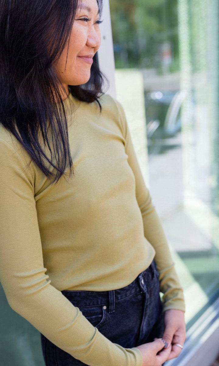 Stillshirt langarm Cléo mit Rollkragen in olive grün kombiniert mit dunkelblauer Jeans vor Fensterscheibe seitlich Blick gerade lächelnd