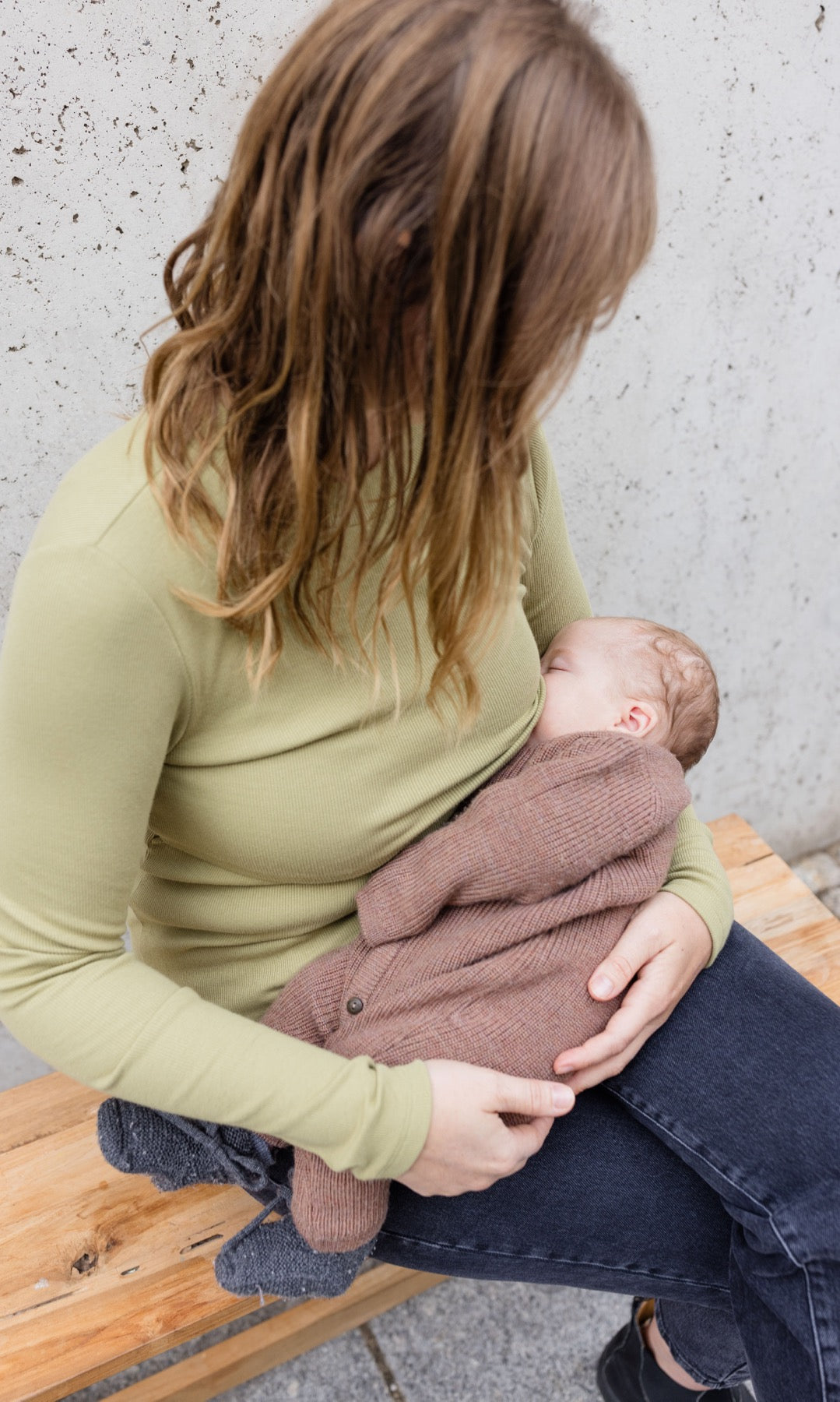 Stillshirt langarm Cléo mit Rollkragen in olive grün kombiniert mit dunkelblauer Jeans sitzend auf Bank mit Baby auf Arm beim Stillen Blick aufs Kind