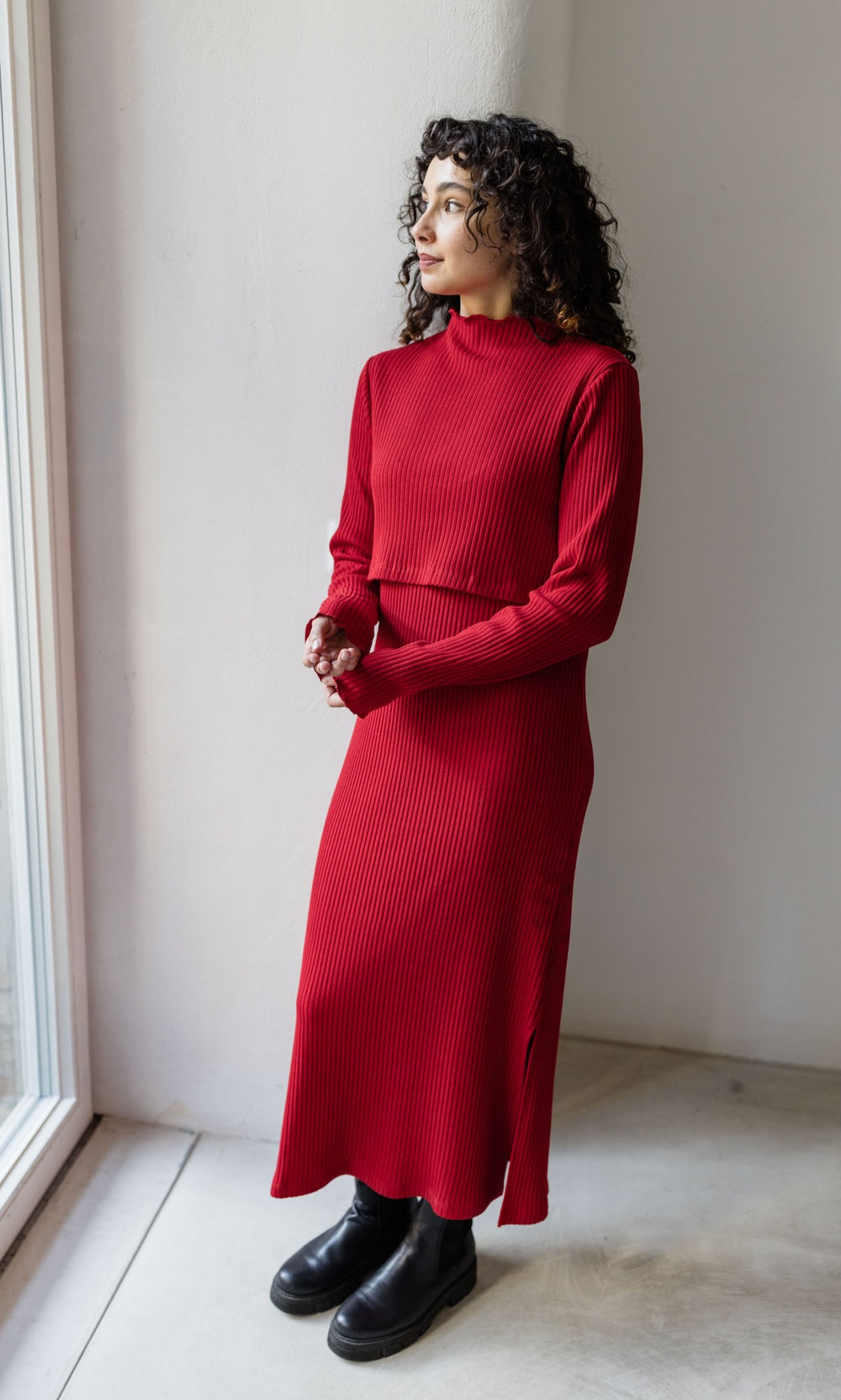 Strickkleid Jil rot lang in langarm mit Stillfunktion Outfit mit schwarzen Stiefeln seitlich stehend vor heller Wand Blick aus Fenster