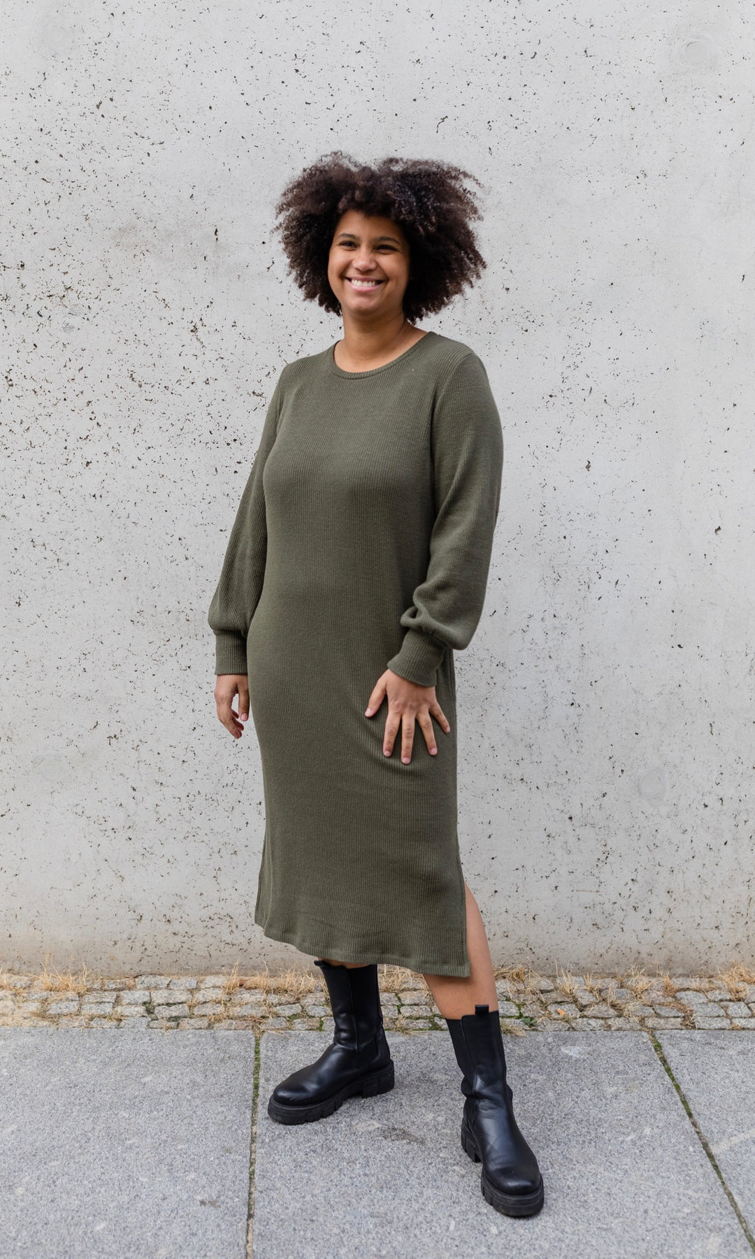 Strickkleid Jules in grün olive mit Stiefeln frontal vor heller Wand