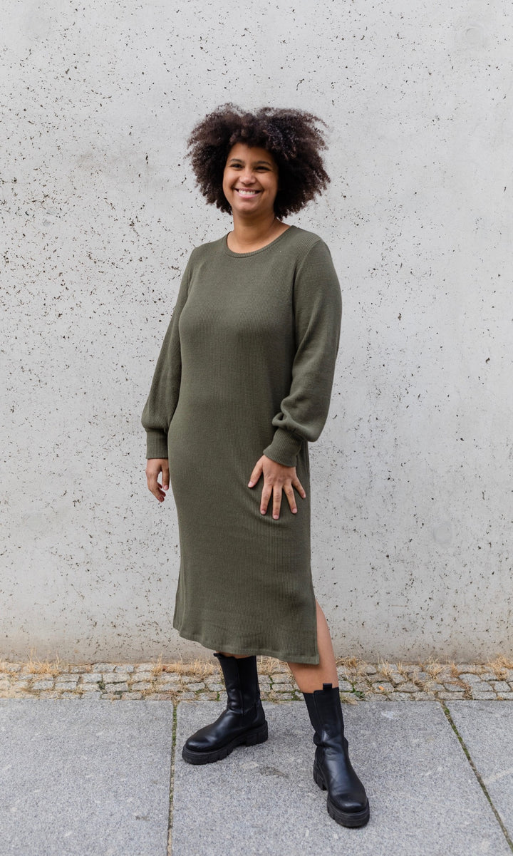 Strickkleid Jules in grün olive mit Stiefeln frontal vor heller Wand