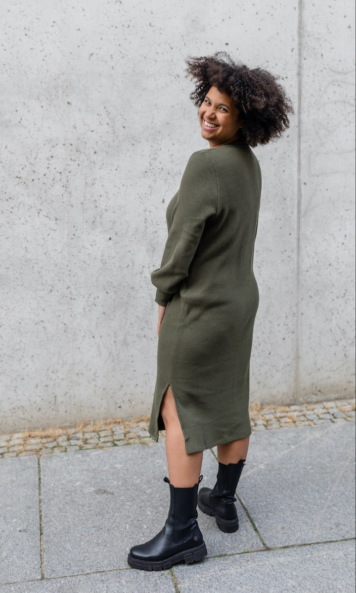 Strickkleid Jules in grün olive mit Stiefeln Ansicht von hinten vor heller Wand