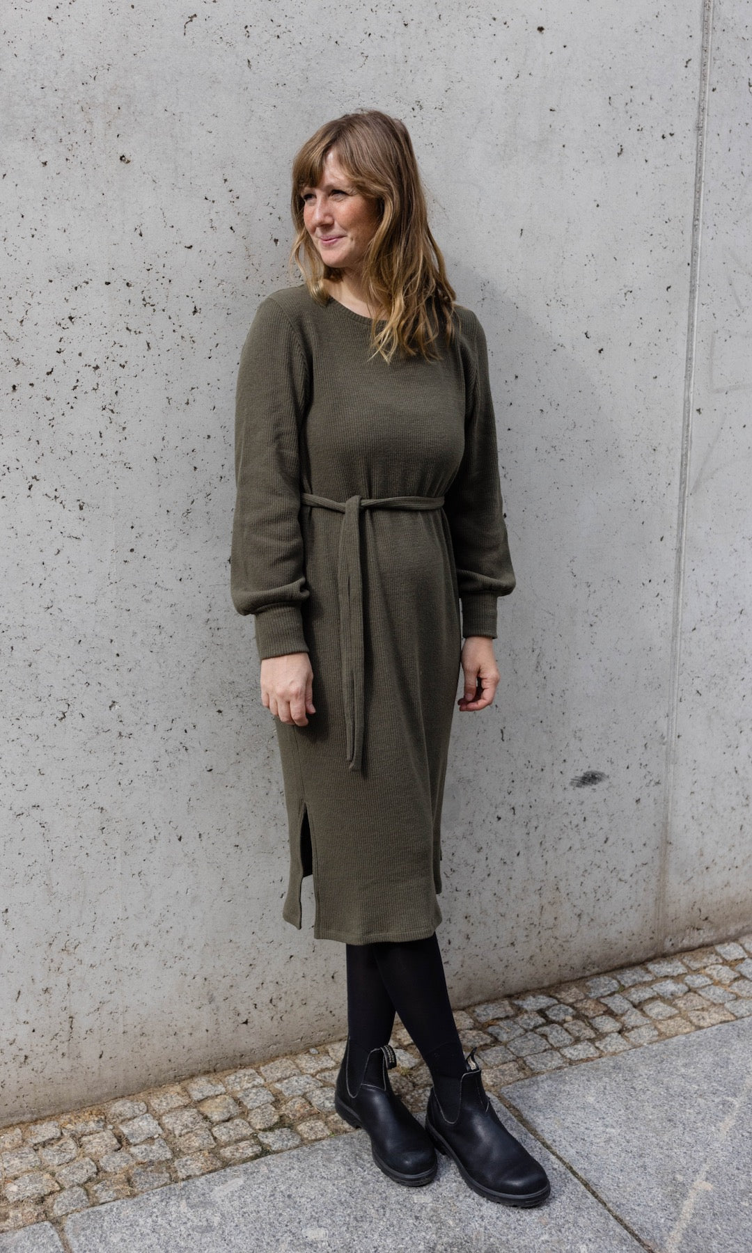 Strickkleid Jules in grün olive mit Stiefeln an heller Wand angelehnt